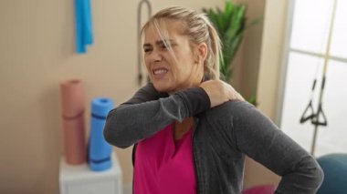 Spor salonunda omuz ağrısı çeken bir kadın arka planda fitness destekleri olan parlak bir egzersiz odasında gerinirken rahatsız edici bir ifade takınıyor..