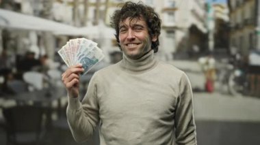 İspanyol asıllı bir adam elinde Polonya zloti banknotları arka planda terası ve kahvecisi olan bir şehir sokağında güvenle gülümsüyor..