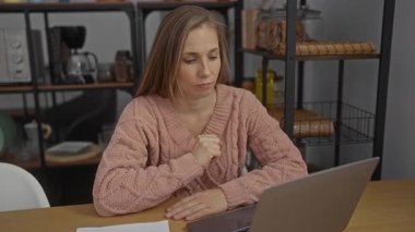 Ofiste dizüstü bilgisayarda çalışan bir kadın kahve makinesi ve arka plandaki raflarla düşünceli bir ifade sergiliyor..