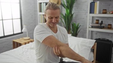 Yatak odasında sarı saçlı beyaz bir adam dirseğini rahatsızlıkla tutarak bir yatakta oturuyor, etrafı ev dekorasyonuyla çevrili ve beyaz bir gömlek giyiyor..