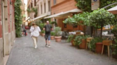 Bulanık görüntü, Roma, İtalya 'da, yeşil ve açık hava kafeteryasında oturan Bokeh' in sakin bir atmosfer yarattığı güzel bir kaldırım taşı caddesinde yürüyen bir erkek ve kadını gösteriyor..