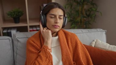 İçeride, kulaklık takmış, rahat bir battaniyeye sarılmış, şık bir oturma odasındaki kanepede müzik dinleyen bir kadın..