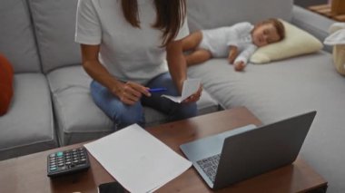Oğlu oturma odasındaki kanepede uyurken evde çalışan bir kadın stresli bir yüz ifadesi sergiliyor ve elinde kalemle evrak işlerine odaklanıyor..