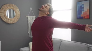 Genç adam, kollarını açmış, rahat bir oturma odasında gülümsüyor. İçinde modern dekorasyon ve parlak doğal ışık var..