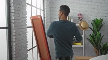 Genç adam şık bir yatak odasında dikilip aynaya bakıyor. Çiçeklerin ve rafların yanında tuğla duvar arkaplanı var. Ev ortamını yansıtıyor ve tefekkür ediyor.