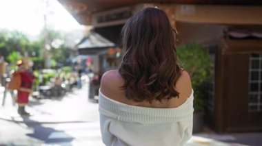 Şehir caddesinde kollarını kavuşturmuş beyaz kazak giymiş, parlak güneş ışığında kameraya gülümseyen bir kadın.