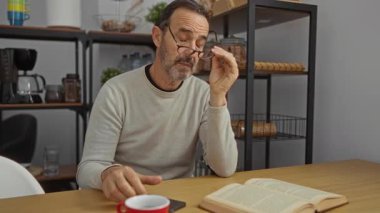 İş yerindeki olgun İspanyol adam, masada kahve ve kitap ile akıllı telefon okumak için bardakları kaldırıyor..