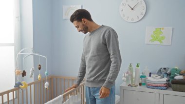 Bebek odasında duran genç adam, oyuncaklarla ve modern ebeveynliği yansıtan temel ihtiyaçlarla çevrili rahat bir evde beşiğe bakıyor.