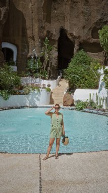 Yeşil elbiseli bir kadın lagomar, lanzarote 'da turkuaz bir havuzun yanında duruyor. Kayalık bir manzarası, yemyeşil bir bahçesi ve arka planda beyaz yıkamalı binaları var..