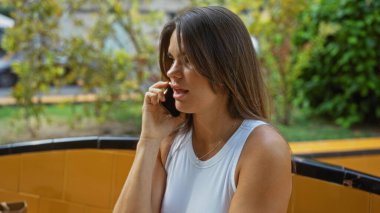 Şehir parkında akıllı telefondan konuşan bir kadın, sakin bir ifade sergiliyor, yeşil, doğal manzara ve güneşli bir günde sarı fayanslı oturma alanı ile çevrili..