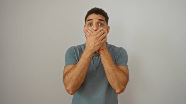 Beyaz arka planda tek başına dikilirken elleriyle ağzını kapatan İspanyol genç adam.