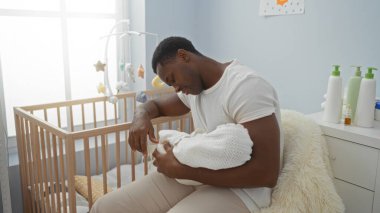 Huzurlu bir yatak odasında oturan genç bir adam, yeni doğmuş bir bebeği beşiğin yanında tutuyor, sıcak ve özenli bir şekilde ev ortamında..