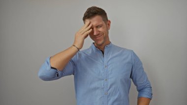 Mavi gömlekli genç adam beyaz arka planda gülümsüyor ve alnını ovuyor.