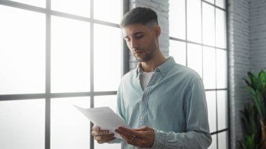 İspanyol bir adam büyük pencereli ve modern dekorlu, konsantrasyon ve profesyonelliği resmeden parlak bir ofis ortamında bir belge okuyor..
