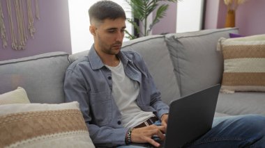 Modern dekorlu, rahat bir oturma odasında dizüstü bilgisayarda çalışan genç İspanyol bir adam şık bir koltukta rahat bir şekilde otururken, odaklanmış ve nişanlı..