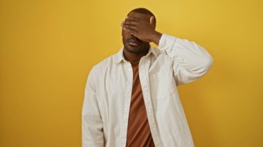 Genç adam gözlerini parlak sarı arka plana karşı eliyle kapatarak çarpıcı, şık ve çağdaş bir görüntü yaratarak çekici ve gizemli varlığını ön plana çıkarıyor..