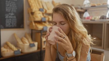 Genç bir kadın, arka planda taze ekmek ve hamur işleri sergilenen sıcak bir fırının içinde kahveyle eğleniyor.