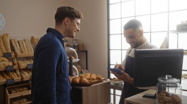 Fırında iki gülümseyen adam ekmek rafları ve pastalarla, bir adam tabletle ödeme yapıyor.