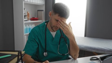 Klinikteki genç İspanyol erkek hemşire masasında stresli görünüyor. Üzerinde yeşil önlükler ve tıbbi ekipmanlar var..