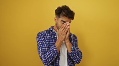 Mavi ekose gömlekli, sakallı genç İspanyol adam sarı arka planda duruyor. Düşünceli bir şekilde ellerini birbirine bastırıyor ve düşünceli duygular aktarıyor..