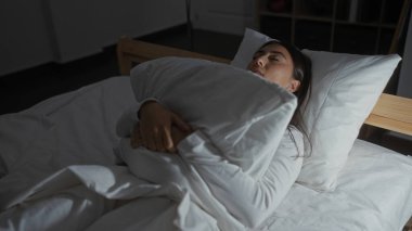 Yatakta elinde yastıkla uyuyan bir kadın loş bir yatak odasında huzurlu bir gece manzarası sergiliyor rahat ve huzurlu bir ambiyansı var.