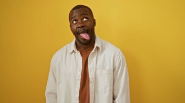 Genç Afrikalı Amerikalı bir adam, dili dışarıda, izole edilmiş sarı arka plan duvarına yaslanmış, komik bir yüz ifadesi takınıyor.