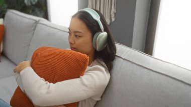 İçeride kulaklıkla müzik dinleyen Çinli genç bir kadın, bir kanepede oturup turuncu yastığa sarılıyor, modern bir oturma odasında..