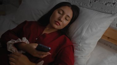 Yatakta uyuyan genç bir kadın, elinde telefon, kırmızı, esmer saçlar, huzurlu bir yüz ifadesi, kapalı bir yatak odası atmosferini çağrıştırıyor..