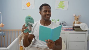 Genç bir adam kitap okuyor ve rahat bir yatak odasında oyuncak tutuyor. Etrafında sıcak bir aile ortamı yaratıyor..