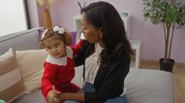 Oturma odasında kızıyla oturan bir kadın sıcak ev ortamında sevgi ve aile bağı gösteriyor. Anne, çocuk, ev içi ve bakım konularını vurguluyor..