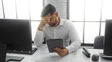 Genç adam modern ofis ortamında iş yerinde elinde tablet tutarken, çevresindeki bilgisayar ve belgelerle işyerindeki gerginliği yansıtıyor..