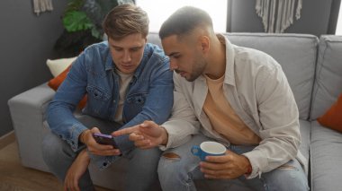 Erkekler rahat bir oturma odasında oturuyorlar, biri akıllı telefonu işaret ediyor, diğeri de kahve bardağını tutuyor, arkadaşça bir etkileşim sergiliyorlar..