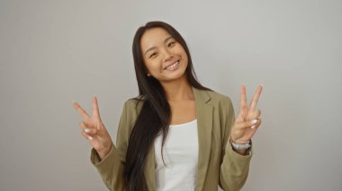 İzole edilmiş beyaz bir arkaplanda poz veren genç Çinli kadın barış işaretlerini iki eliyle gösteriyor, pozitifliği ve mutluluğu ifade ediyor.
