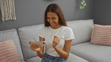 Genç bir kadın rahat bir oturma odasında telefonuna bakıp gülümsüyor. Evin içinde modern dekorasyon ve doğal ışık filtresi olan neşeli bir iç mekan yaratıyor..