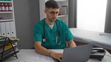 Tıbbi önlüklü İspanyol genç adam, klinik odasında dizüstü bilgisayarda çalışıyor. Dosyalar ve steteskop içeriden görülüyor. Odaklanmış profesyonel bir ortam olduğunu gösteriyor.