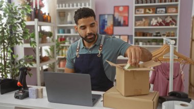 Delikanlı, ev dekorasyonu ambalaj ürünleri satan bir mağazada çalışıyor. Etrafı bitki, resim ve dekoratif eşyalarla dolu raflarla çevrili.