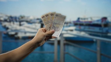 Canlı bir sahil limanının arka planına karşı elinde Türk lirası banknotları tutan bir adam. Tekneler ve sakin su ile, finans ve seyahat öyküsü yaratıyor..