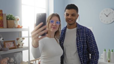 Çift sıcak yatak odasında kadın ve adamla selfie çekiyor. Doldurulmuş hayvanlar ve dekorla çevrili beşiğin yanında gülümsüyor. Parlak, modern ev içi mekanda..