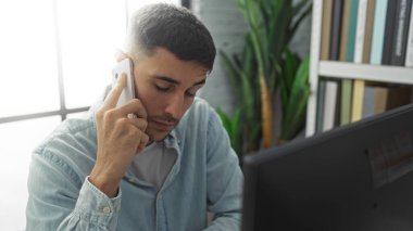 Ofisteki genç adam akıllı telefondan konuşuyor, etrafı kitaplar ve bitkilerle çevrili, profesyonel ve odaklanmış bir atmosferi çağdaş bir iç mekan ortamında taşıyor..