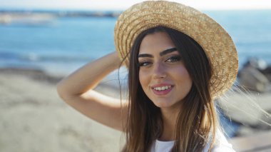 Güneşli bir günde deniz kenarındaki gezinti alanında hasır şapka takan, çekici esmer bir kadın, neşeli yaz aylarında açık hava kumsal manzarasının keyfini çıkarıyor..