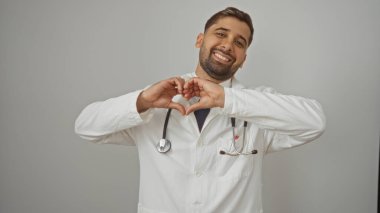 Yakışıklı genç İspanyol doktor beyaz arka planda içten bir gülümsemeyle elleri laboratuvar önlüğü ve steteskop ile kalp sembolü yaparak dostça yaklaşılabilir bir sahne yaratıyor.