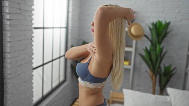 Yatak odasının yanındaki sarı saçlı kadın iç çamaşırlarını bitkilerle ve büyük pencerelerle düzeltiyor..