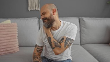 Sakallı ve dövmeli yakışıklı bir adam modern bir oturma odasında boynu ağrıyan şık bir dekorasyonla evde rahat bir koltukta otururken rahatsızlığını dile getiriyor.