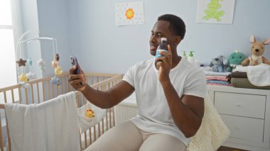 Yatak odasında beşiği ve oyuncaklarla çevrili bir şekilde bebek şişesini tutarken selfie çeken yakışıklı bir adam sıcak bir yuva kurarken neşeli bir anı yakalıyor..
