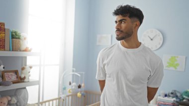 Yakışıklı bir adam beşiği olan parlak bir bebek odasında duruyor, oyuncaklar ve pastel süslemelerle çevrili, sakin ve besleyici bir atmosfer sunuyor..