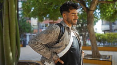 Sakallı İspanyol bir adam şehir parkında duruyor, şık bir ceket ve sırt çantası giyiyor, ağaçlarla çevrili, açık havada şehir atmosferinin tadını çıkarıyor..