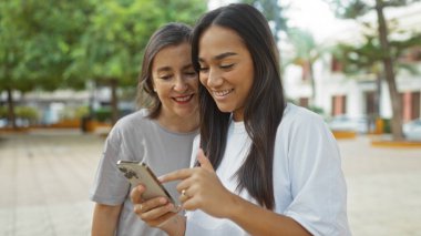 Şehir parkında başka bir kadına akıllı telefon gösterirken gülümseyen bir kadın aile sevgisi ve arkadaşlığını resmediyor..
