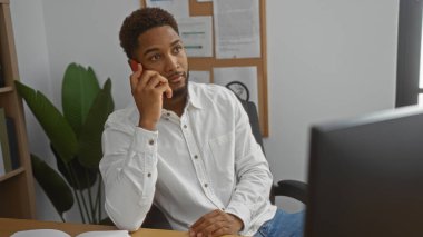 Yakışıklı, Afro-Amerikalı bir adam. Ofis ortamında telefonla konuşuyor. Evrakları ve arka planda bir bitki var.