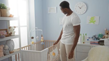 Genç adam sıcak bir yatak odasında, içi doldurulmuş hayvanlar ve hafif aydınlatmalarla çevrili şefkat ve sıcaklıkla bir bebek beşiğinin yanında duruyor, davetkar bir ev atmosferi yaratıyor.