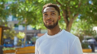 Güneşli bir şehir parkında arka planda ağaçlar ve binalar olan yakışıklı genç adam beyaz bir tişört giyiyor ve düşünceli bir şekilde gülümsüyor.